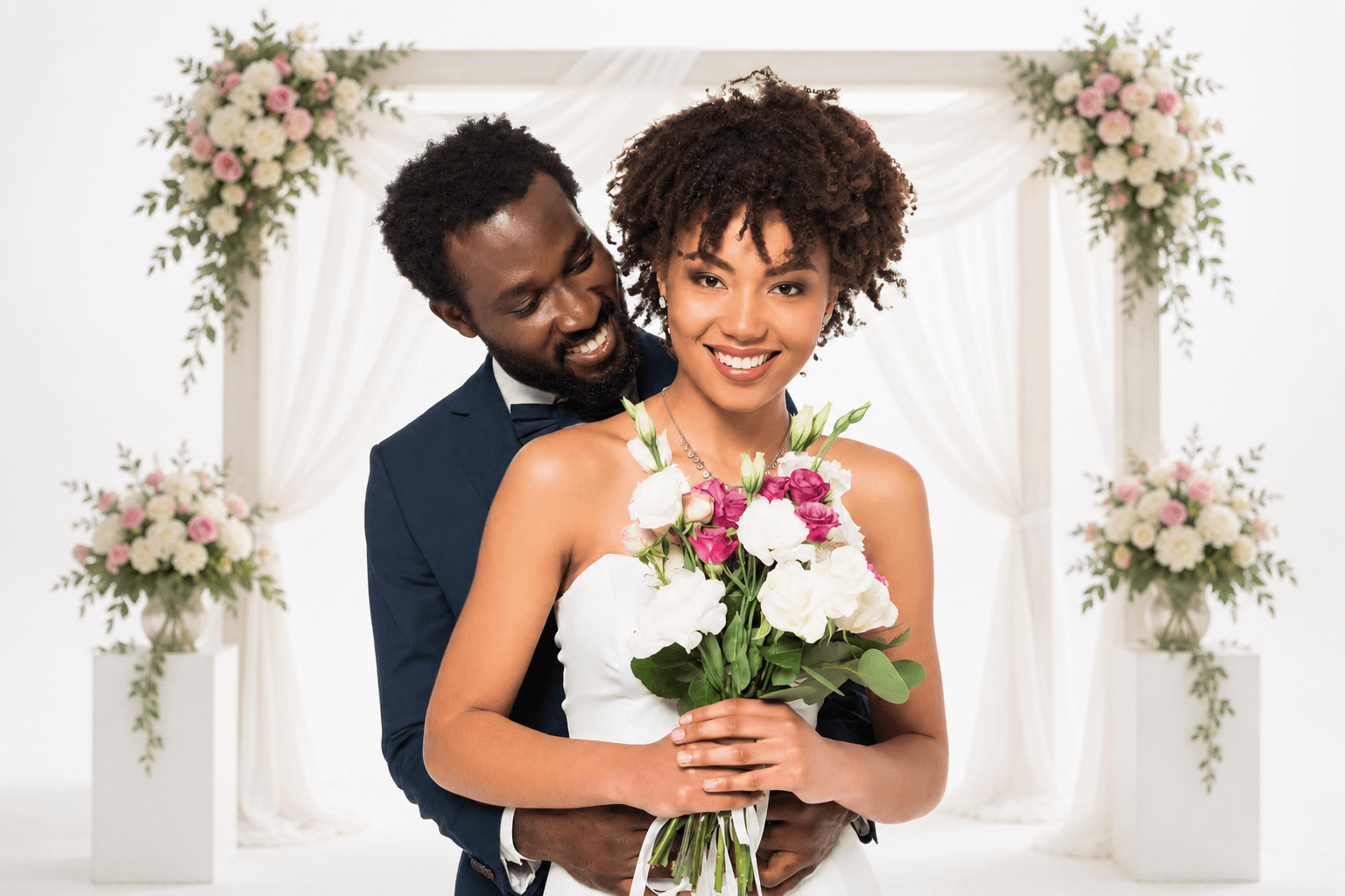African-American bride holding a bouquet, embraced by her groom beneath a floral wedding arch